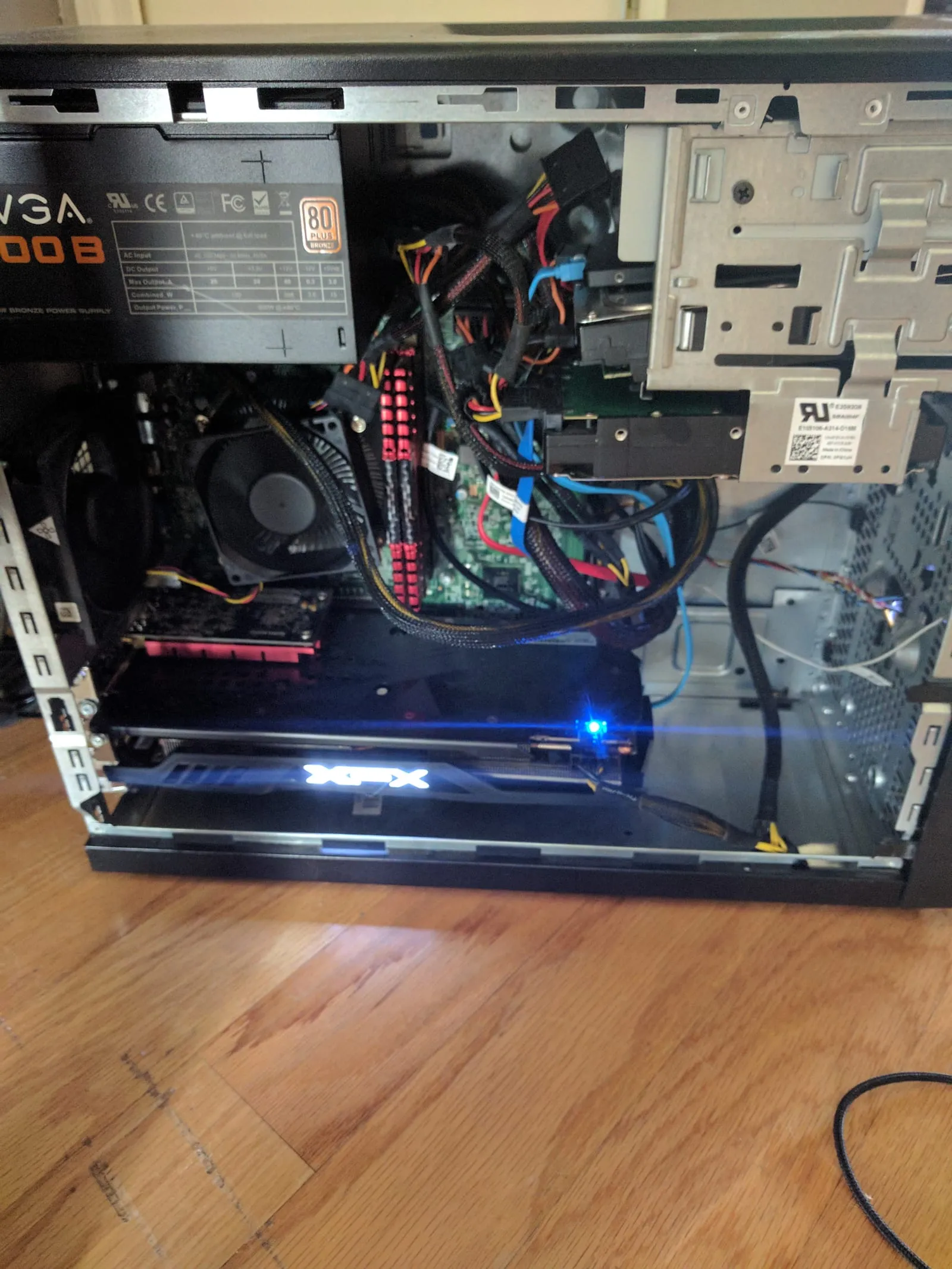 An open desktop PC case on a hardwood floor with a box fan strapped to the side blowing onto the graphics card