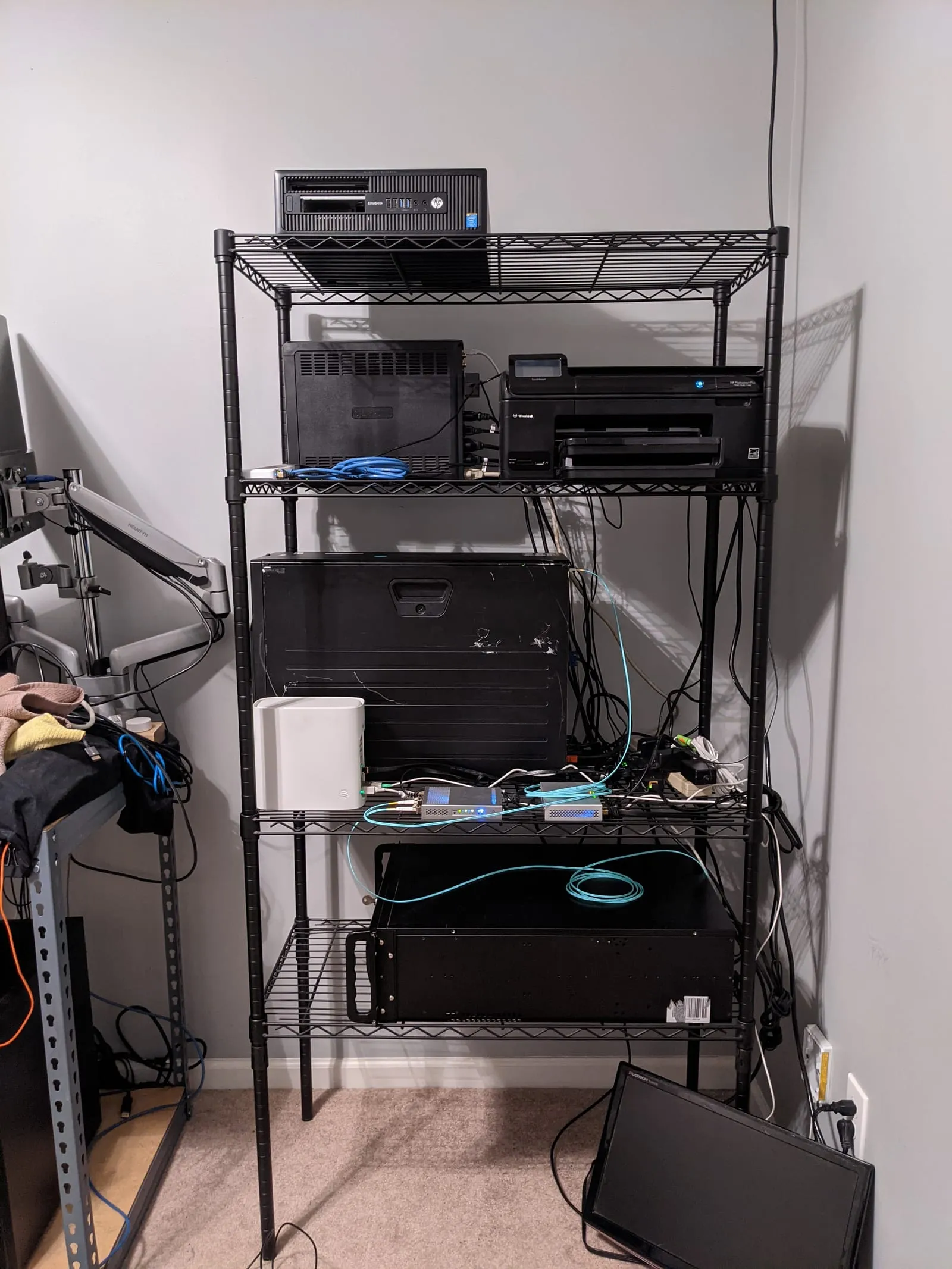 A black wire shelving unit holding an HP small-form-factor PC, a rackmount chassis, a printer, and a tangle of blue network cables