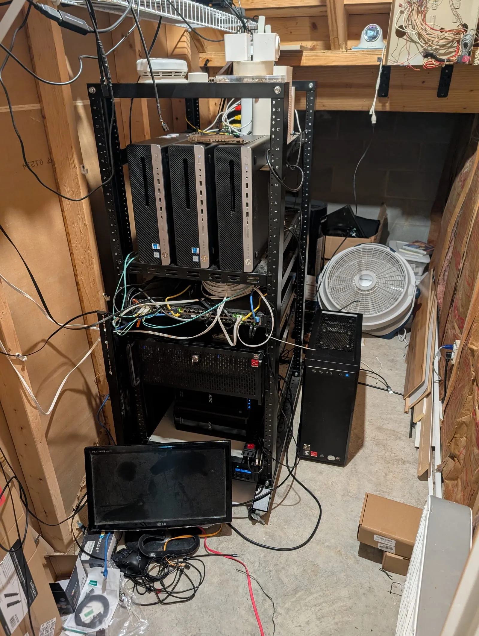 A two-post open server rack in a basement holding three stacked HP small-form-factor PCs, rackmount chassis and patch cables, with a box fan beside it and a concrete block wall behind